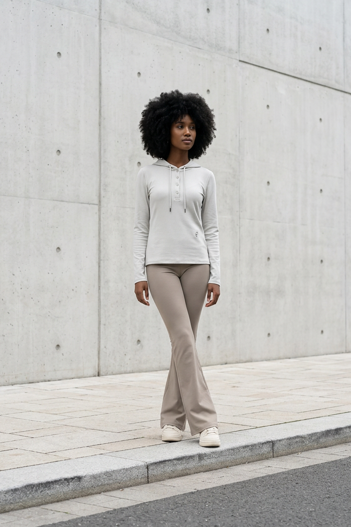 A woman with an afro stands on a sidewalk in front of a large concrete wall, wearing a light beige long-sleeve hoodie and matching beige flared pants with white sneakers.