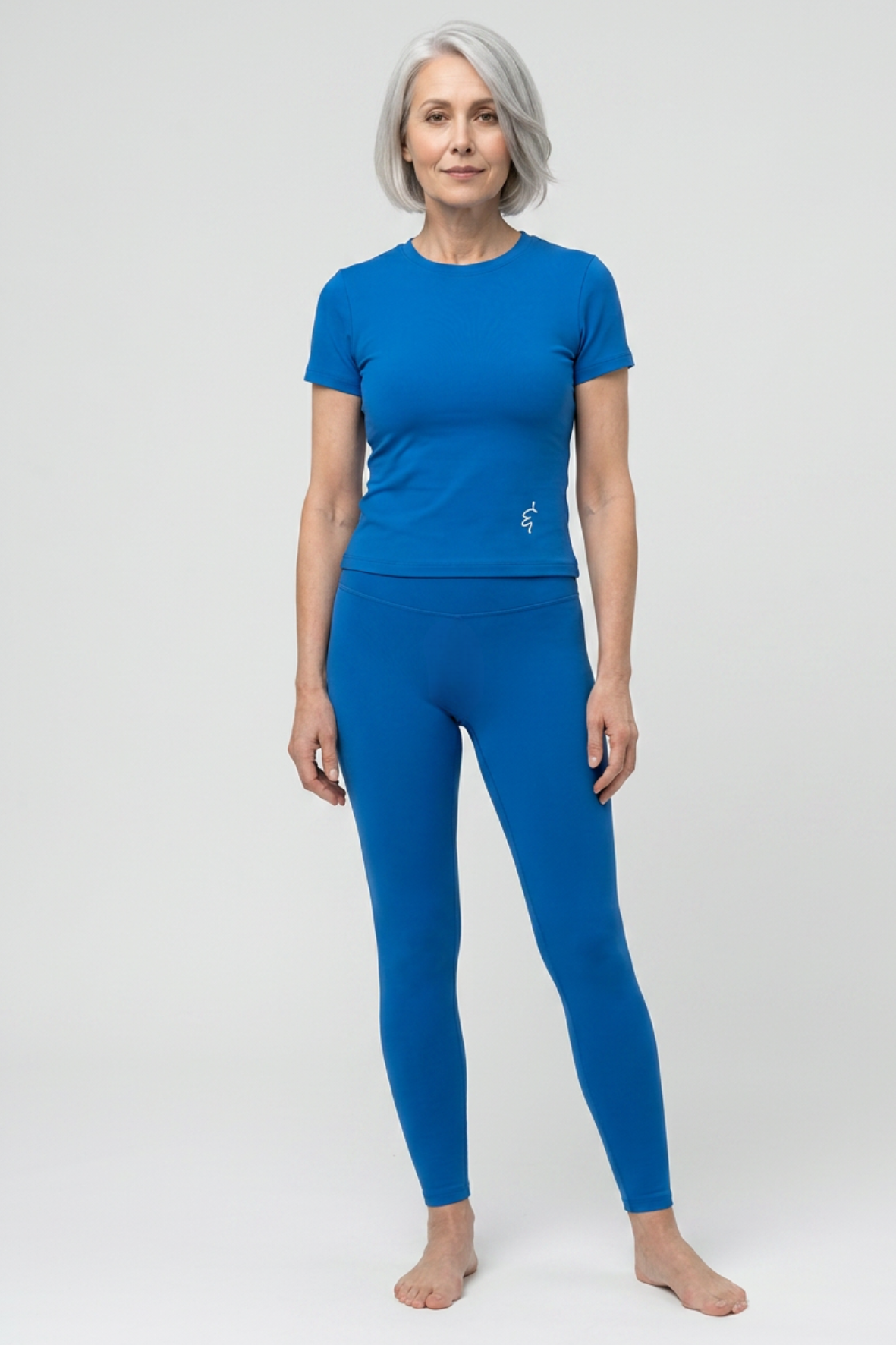A woman with short gray hair stands barefoot against a light gray studio background, wearing a bright blue short-sleeve crew-neck top and matching blue leggings.