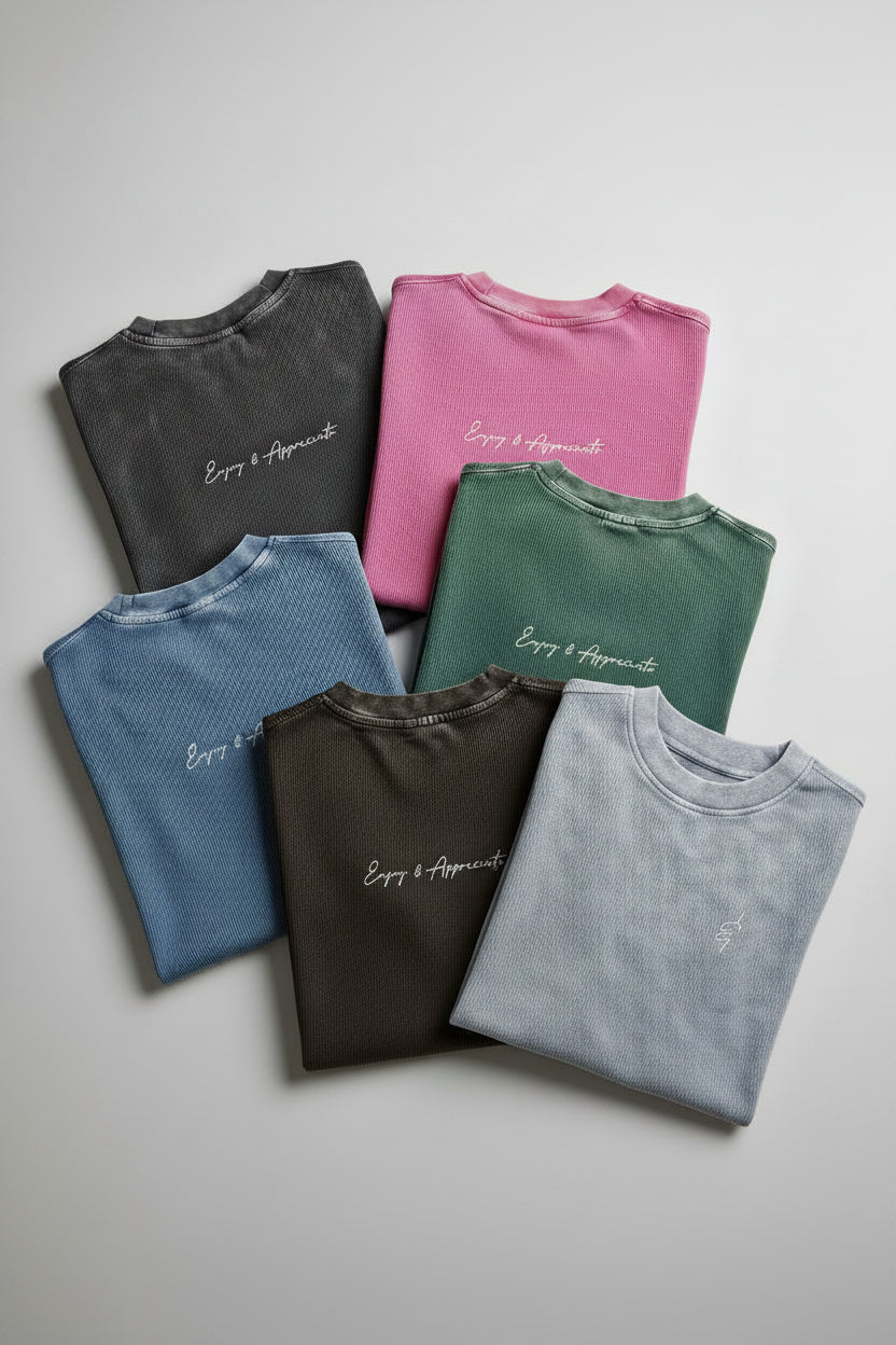 Six folded short-sleeve t-shirts in assorted muted colors are arranged in a fan shape on a beige linen fabric background. The shirts include charcoal, pink, sage green, dusty blue, black, and heather gray, each with a small white script logo on the chest reading 'Every & Everything'.