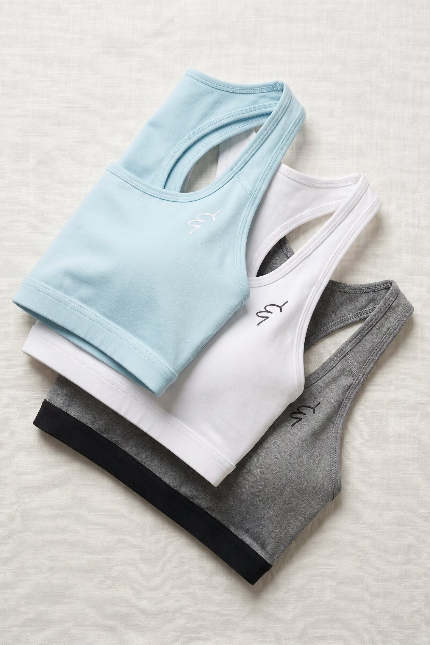 Three racerback sports bras in light blue, white, and heather gray, arranged in a fan-like stack on a light beige fabric background. Each bra features a small embroidered logo on the upper chest.