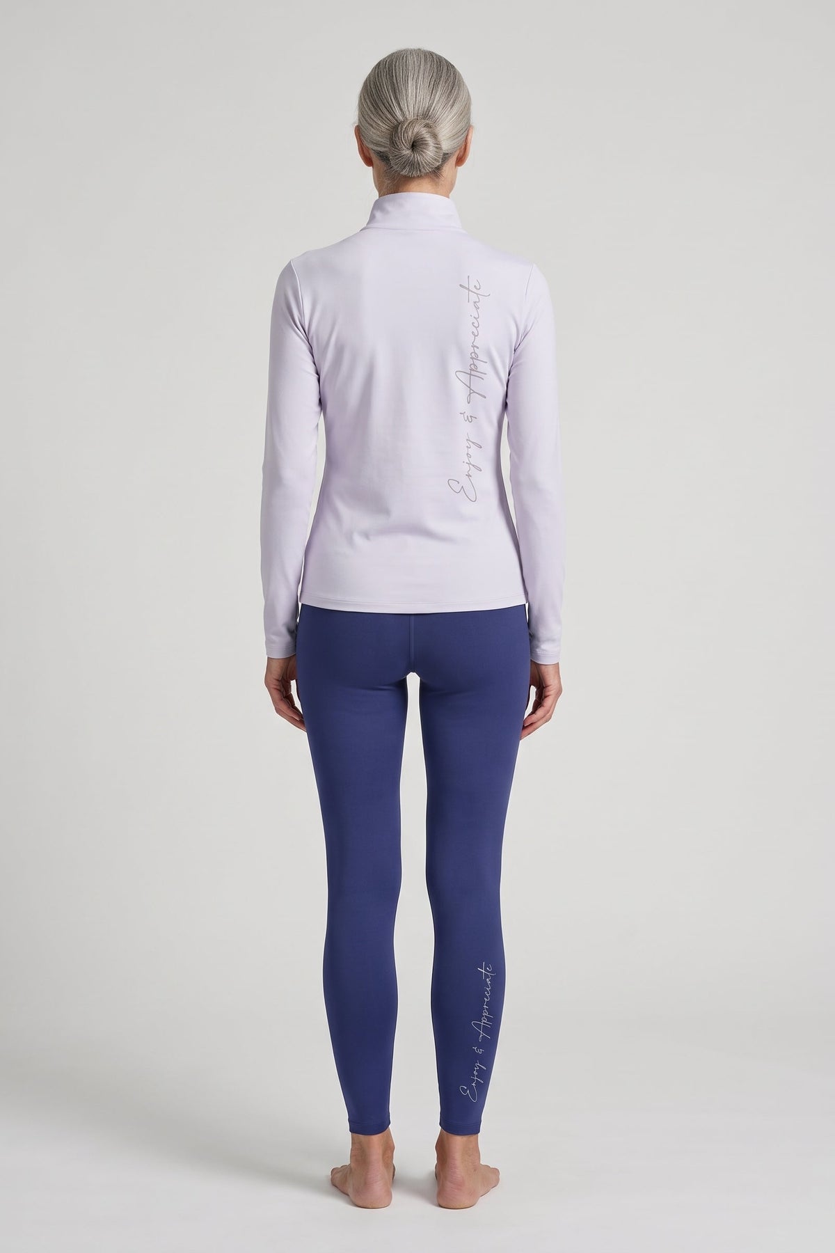 A woman stands barefoot against a light gray studio background, viewed from the back. She is wearing a pale lavender long-sleeve athletic top and dark blue leggings, each with vertical script branding down the back and leg respectively.