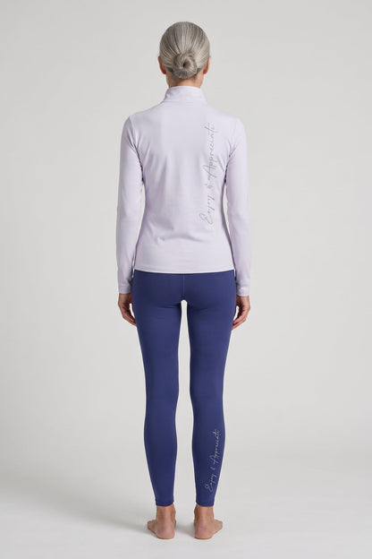 A woman stands barefoot against a light gray studio background, viewed from the back. She is wearing a pale lavender long-sleeve athletic top and dark blue leggings, each with vertical script branding down the back and leg respectively.