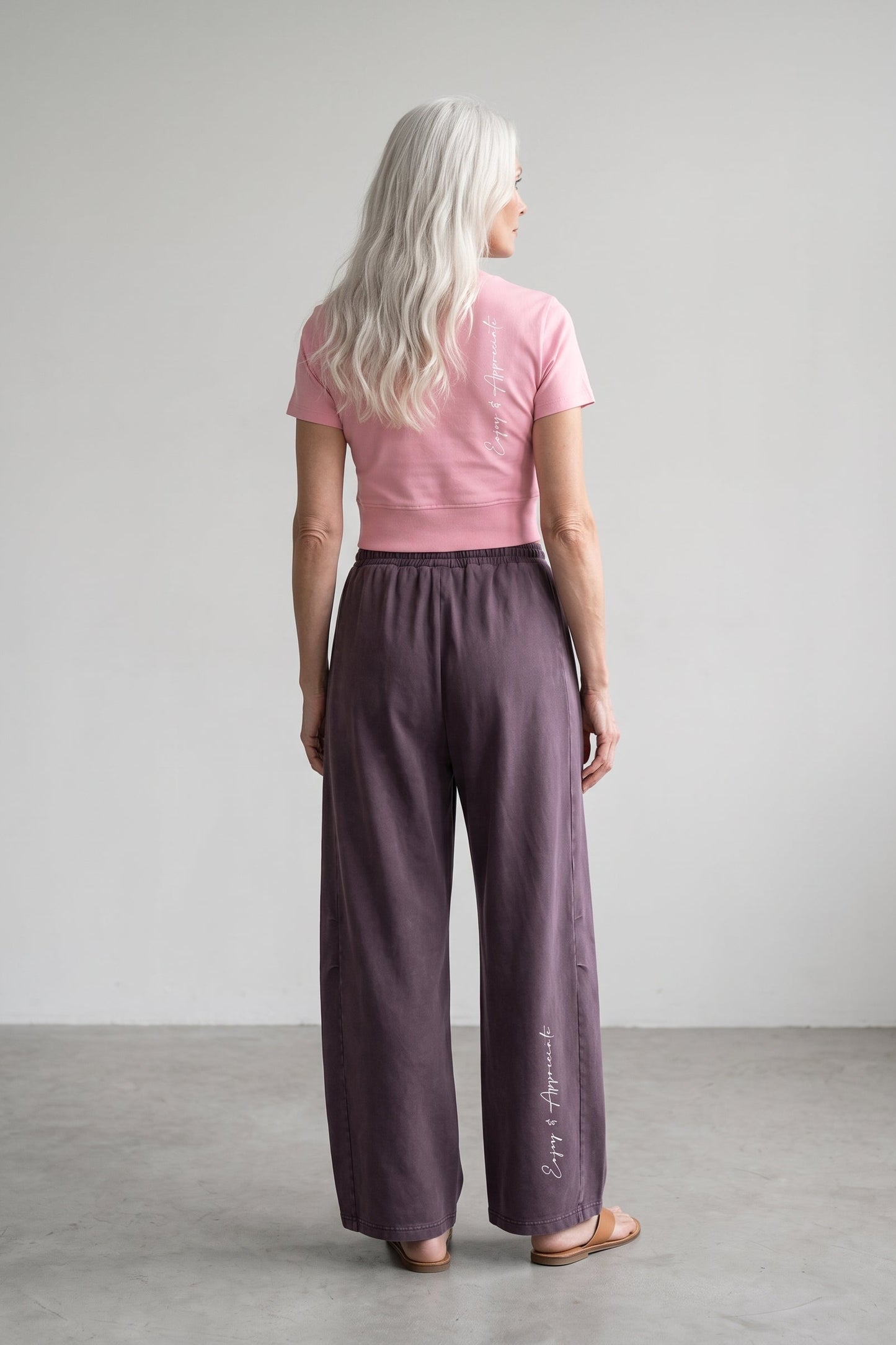 A woman with long platinum blonde hair is shown from behind wearing wide-leg mauve-purple trousers and a pink t-shirt, standing in a minimalist studio setting.