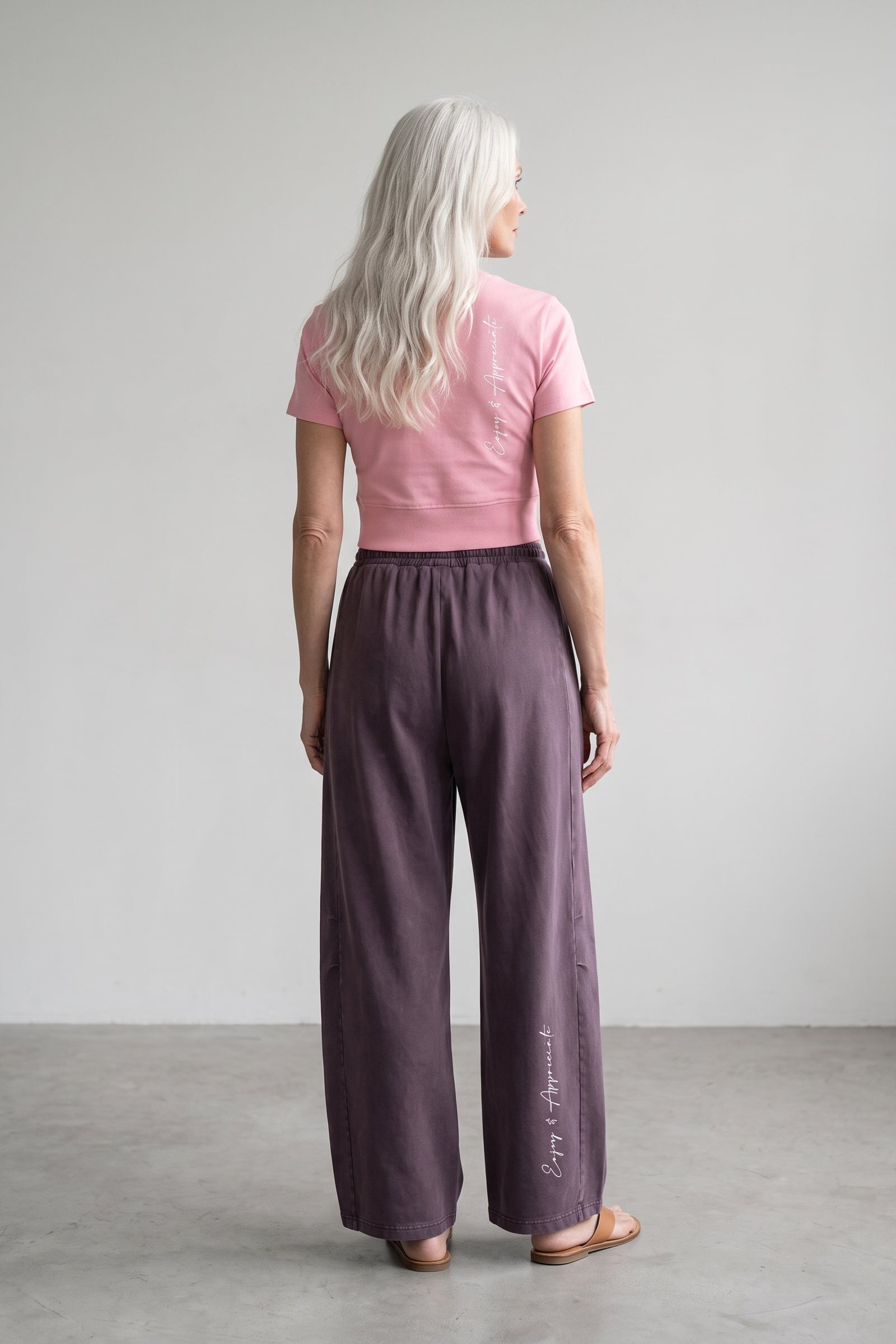 A woman with long platinum blonde hair is shown from behind wearing wide-leg mauve-purple trousers and a pink t-shirt, standing in a minimalist studio setting.