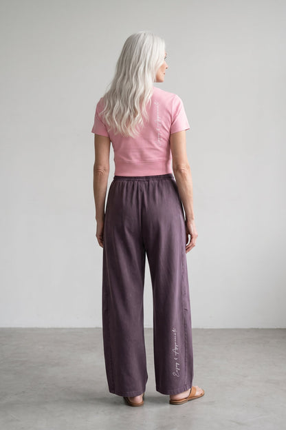 A woman with long platinum blonde hair is shown from behind wearing wide-leg mauve-purple trousers and a pink t-shirt, standing in a minimalist studio setting.