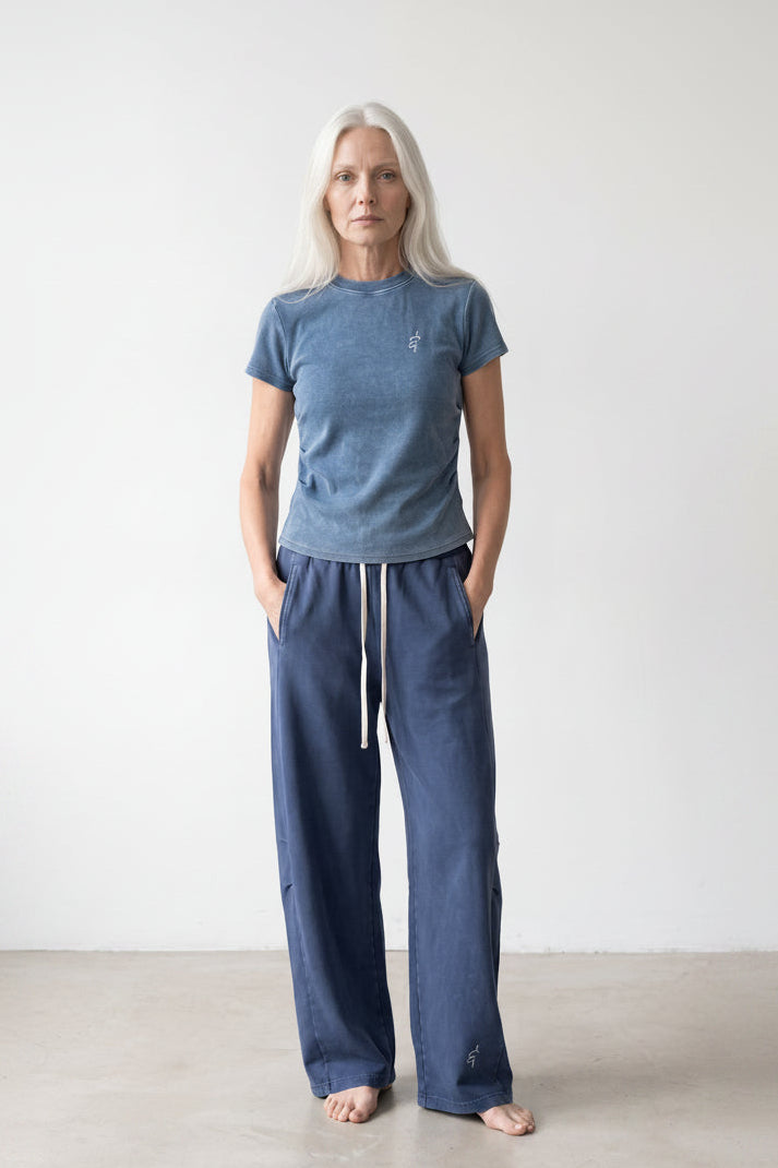 A woman with long white hair stands barefoot in a minimalist studio setting, wearing a heathered blue short-sleeve t-shirt and matching blue wide-leg drawstring pants.
