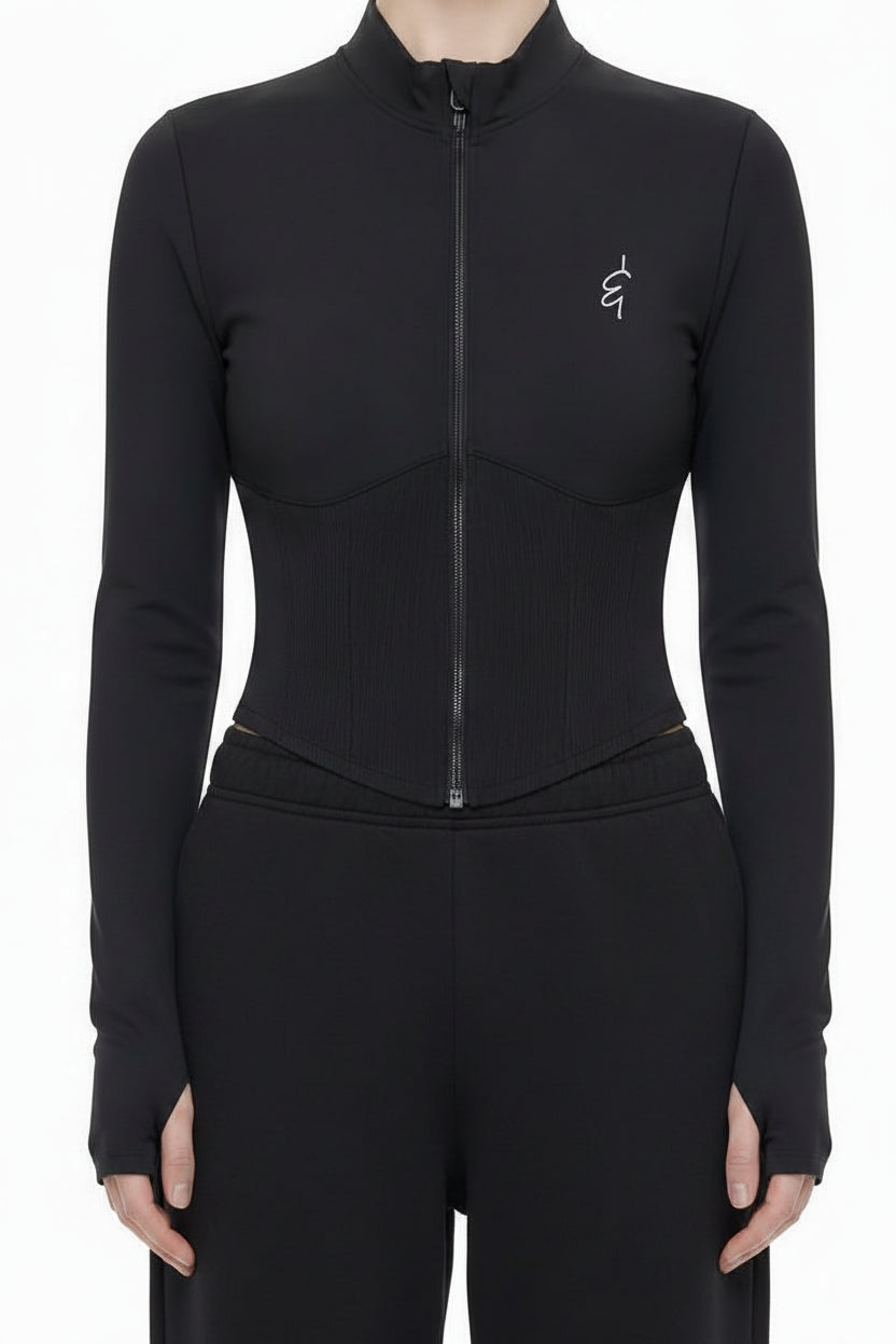 A model stands against a white studio background wearing a black long-sleeve zip-front bodysuit with a small embroidered logo on the left chest, paired with light gray wide-leg sweatpants and white sneakers.