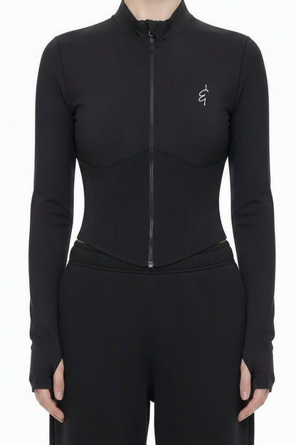 A model stands against a white studio background wearing a black long-sleeve zip-front bodysuit with a small embroidered logo on the left chest, paired with light gray wide-leg sweatpants and white sneakers.