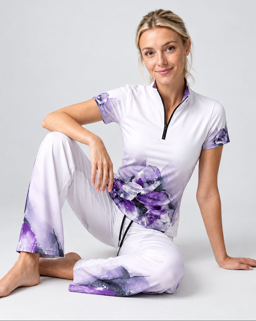 A woman models a matching scrub set consisting of a short-sleeve half-zip top and coordinating pants. The scrubs are primarily white with a purple watercolor floral pattern on the lower torso and sleeve cuffs, and a black zipper accent.
