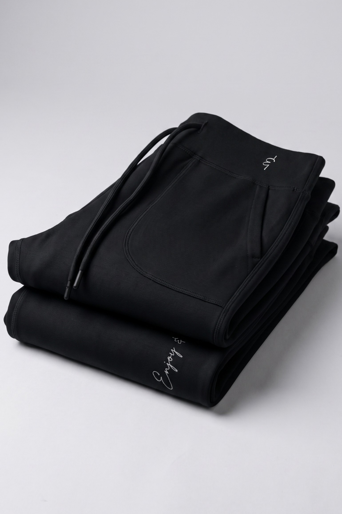 A pair of folded black sweatpants with white embroidered script logos on the side, displayed on a light gray studio background.