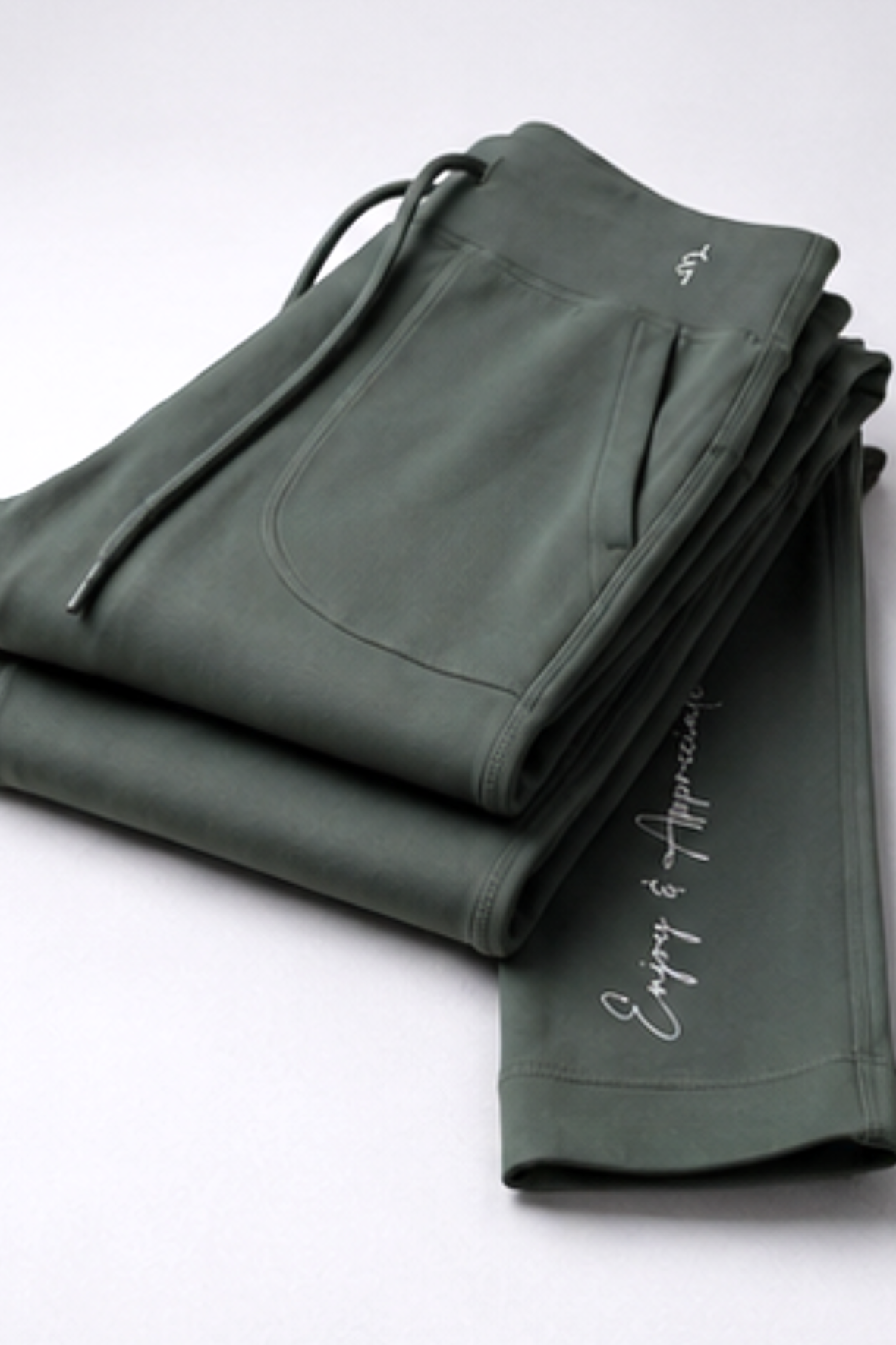 A pair of folded dark olive green sweatpants with white script embroidery on the leg and a drawstring waist, displayed on a clean white background.