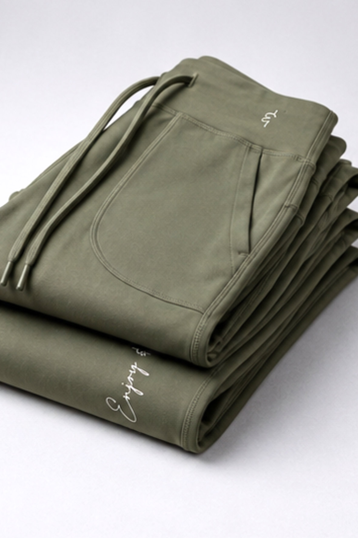 A stack of olive green folded sweatpants on a light gray background. The sweatpants feature a drawstring waist, a front kangaroo pocket, and white script embroidery on the leg.