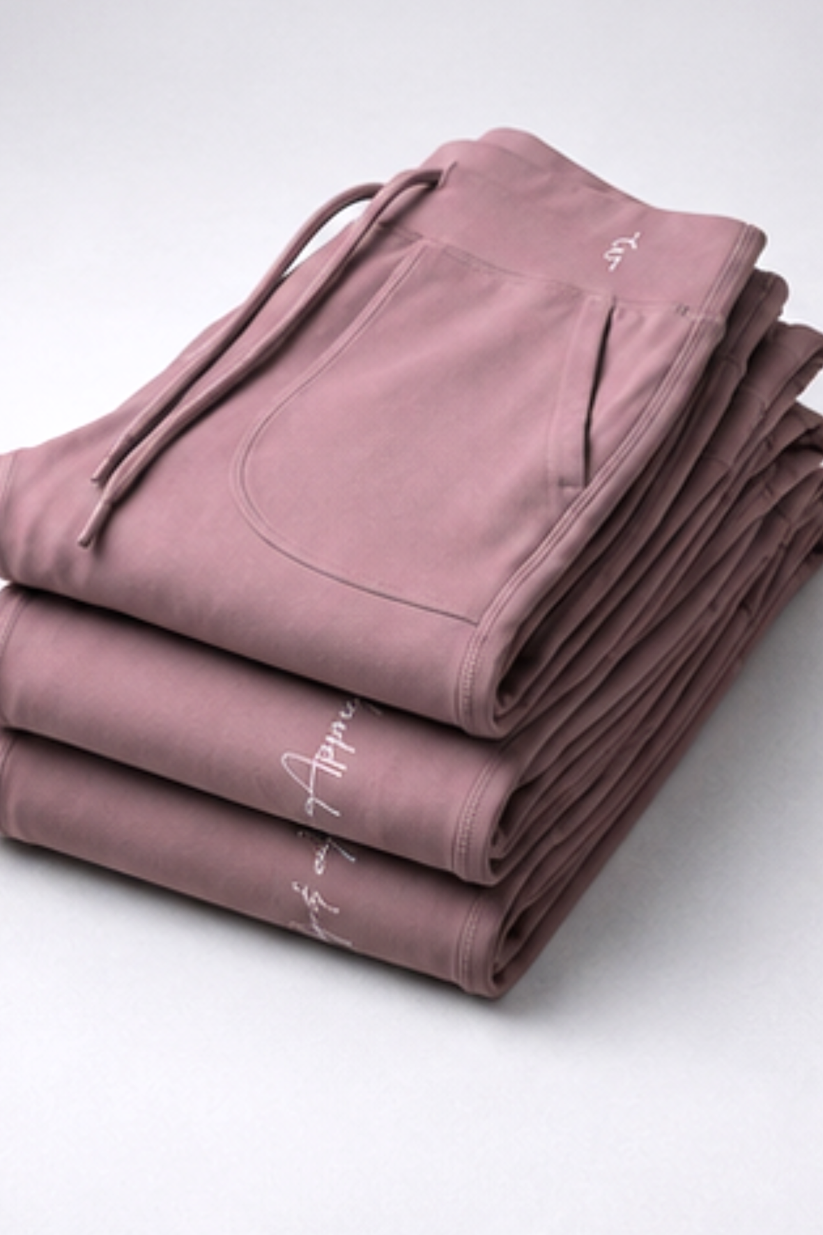 A stack of folded dusty rose drawstring sweatpants with white script embroidery on the side, photographed on a clean light gray studio background.