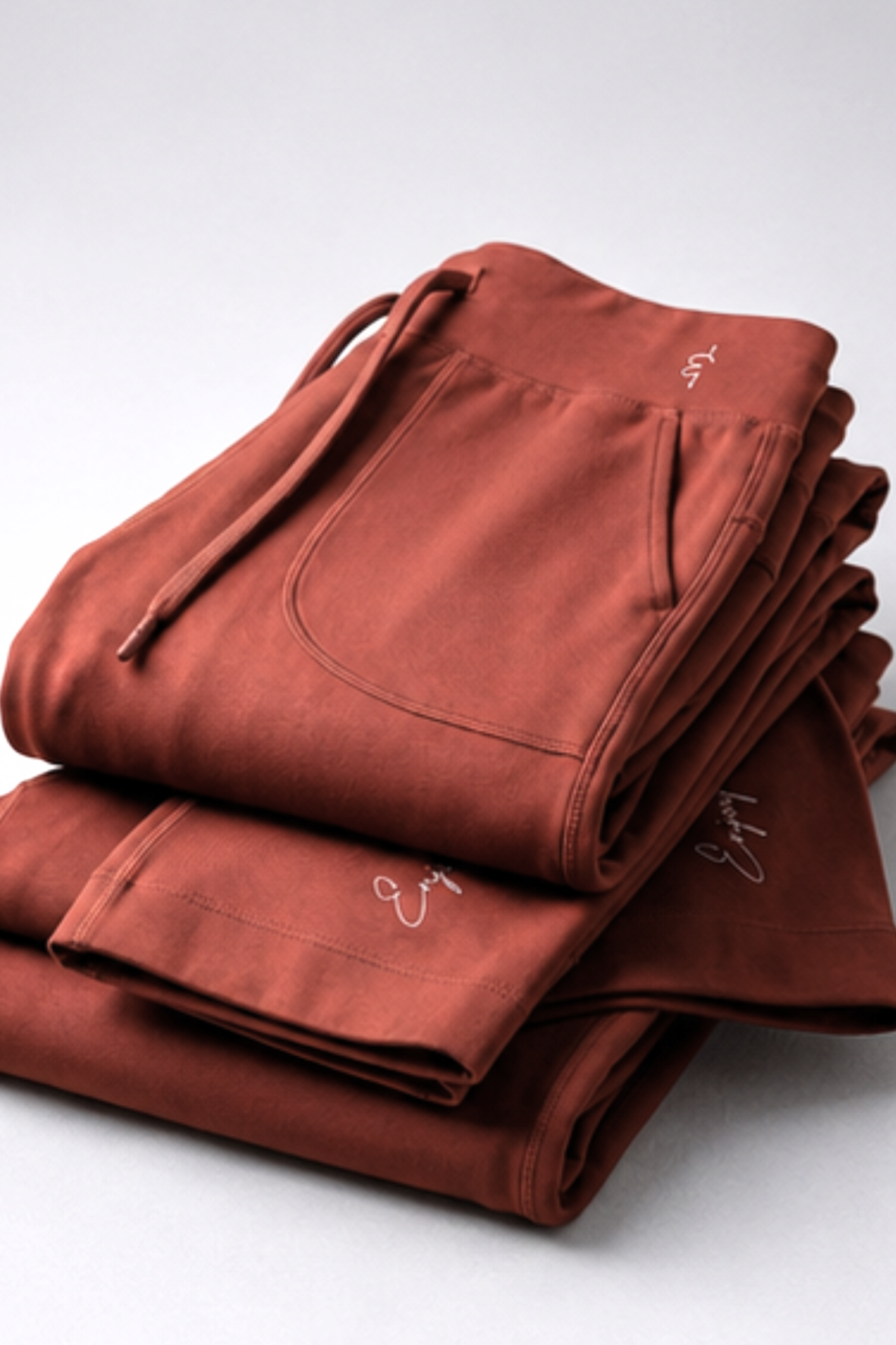 A stack of folded terracotta-colored sweatpants with drawstrings and side pockets, photographed on a clean light gray studio background.