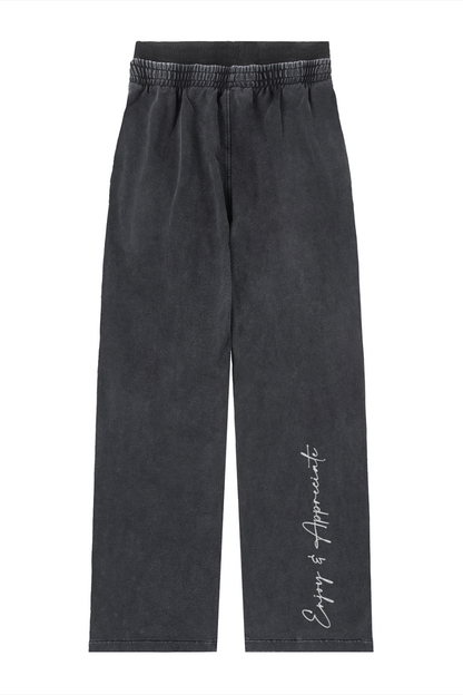 A pair of dark gray washed wide-leg pants with an elastic waistband and a white script logo reading 'Enjoy & Appreciate' printed down the left leg, shown against a plain white background.