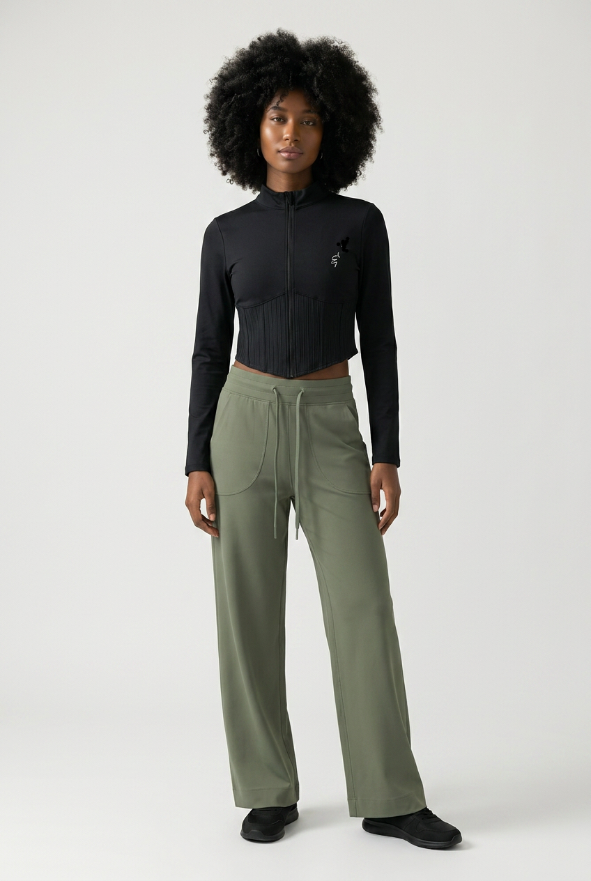 A model with an afro hairstyle stands against a light gray studio background wearing a black long-sleeve cropped zip-up top and olive green wide-leg drawstring pants.