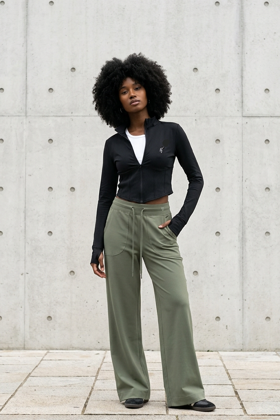 A woman with an afro stands on a sidewalk in front of a concrete wall, wearing a black long-sleeve cropped top over a white undershirt and olive green wide-leg trousers.