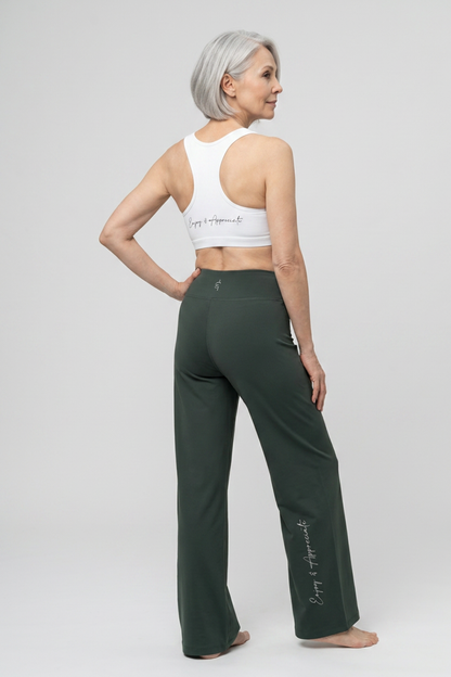 An older woman with short gray hair is shown from the back wearing dark forest green wide-leg yoga pants and a white racerback sports bra. Both garments feature the script brand name 'Crazy Appropriate' printed on them.