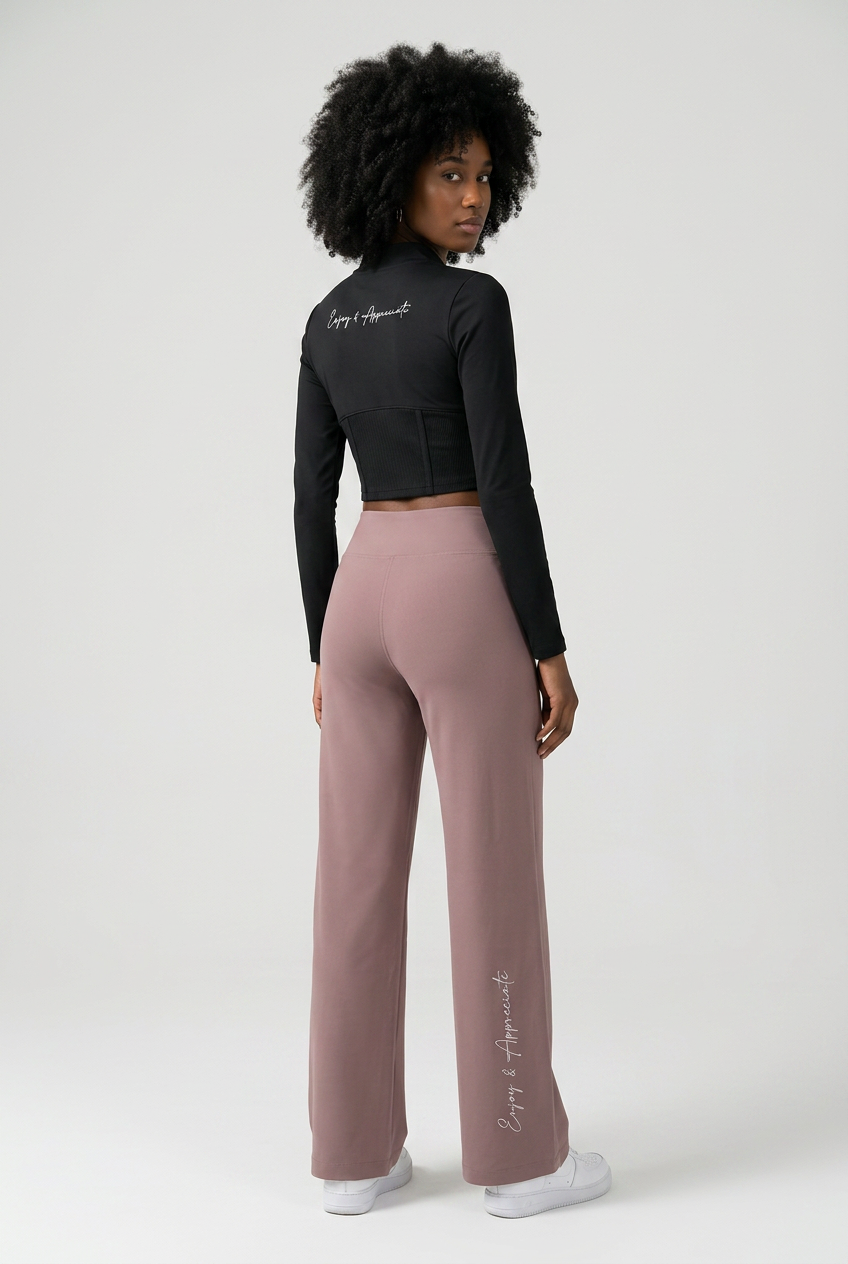 A woman is shown from the back wearing a black long-sleeve crop top and dusty rose wide-leg pants with white sneakers, set against a plain light gray studio background.