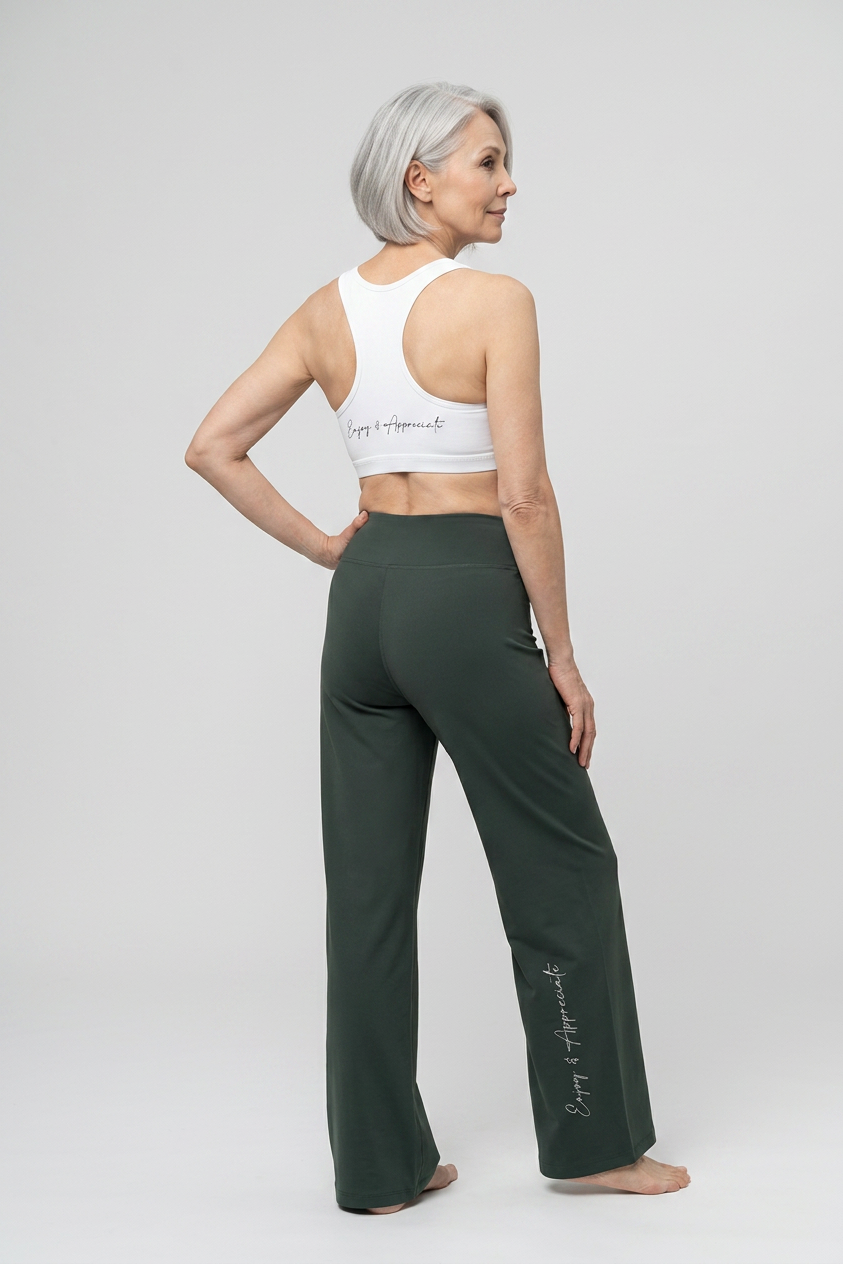 A woman with short gray hair is shown from the back wearing dark green wide-leg yoga pants and a white sports bra with script text, standing against a light gray studio background.