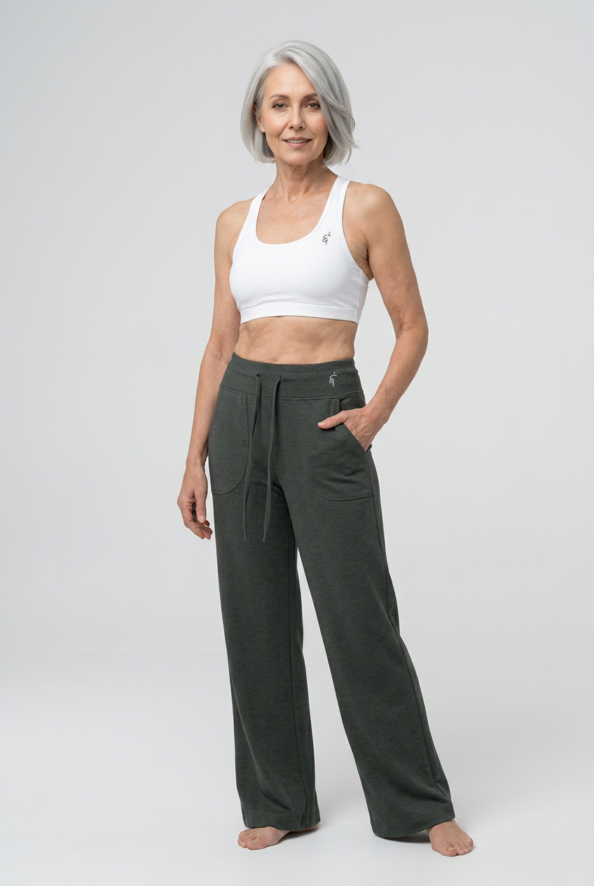 A woman with short gray hair stands barefoot against a light gray studio background, wearing a white sports bra and dark olive green wide-leg lounge pants with a drawstring waist and side pockets.