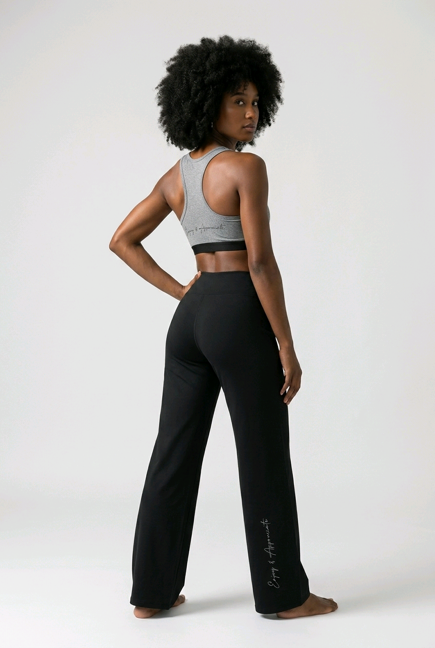 A woman stands barefoot against a light gray studio background, photographed from the back and side. She is wearing black wide-leg yoga pants with a small white script logo near the lower left leg and a gray sports bra.