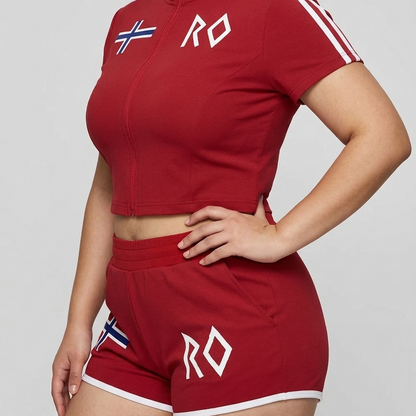 Model wearing a red women's cropped full-zip activewear top and matching athletic shorts with white trim, Norwegian flag detail and RO logo.