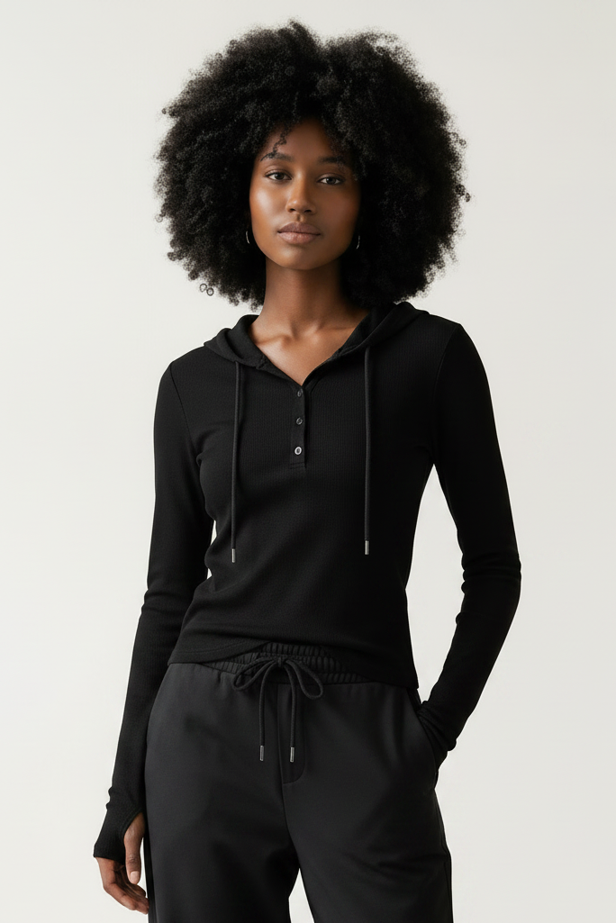 A woman with a large black afro stands in a minimalist room with light wood floors and a white wall, wearing a dark gray long-sleeve zip-up hoodie, black wide-leg pants, and white sneakers.