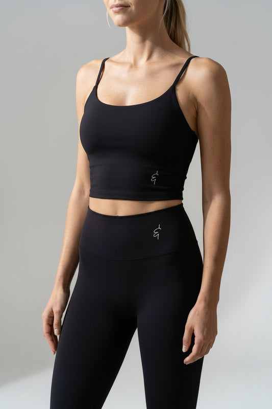 A woman modeling a matching black activewear set consisting of a scoop-neck crop top and high-waisted leggings, both featuring a small white stylized logo on the front.