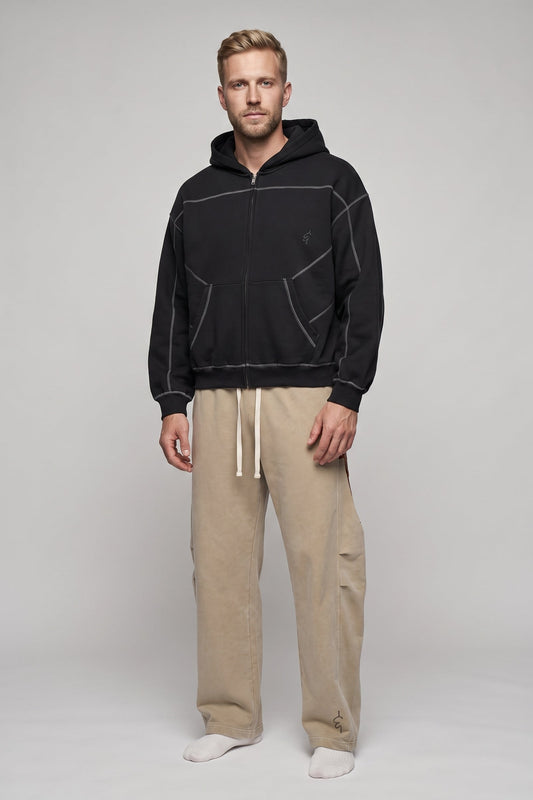 A man modeling a black zip-up hoodie with contrasting light gray stitching and beige drawstring pants against a light gray studio background.