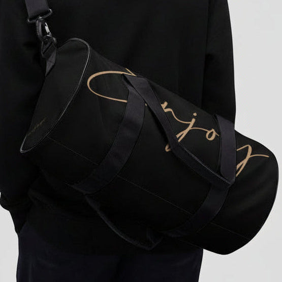 Black cylindrical gym duffel bag with gold cursive lettering and black straps for fitness and travel use