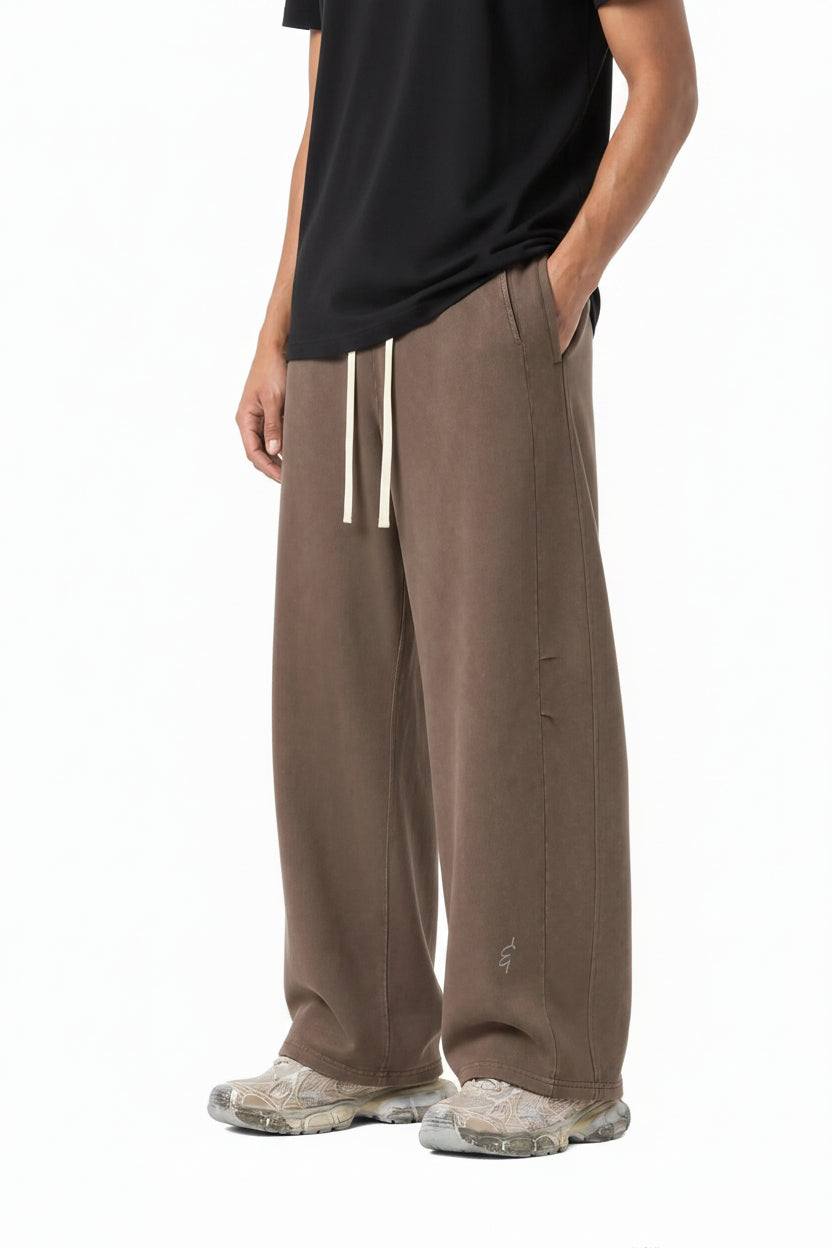 A person modeling a pair of loose, wide-leg brown sweatpants with a white drawstring, shown from the waist down against a plain white background.
