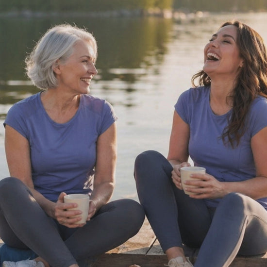 Two women wearing blue t-shirts sitting on a deck holding cups with water and mountains in the background.