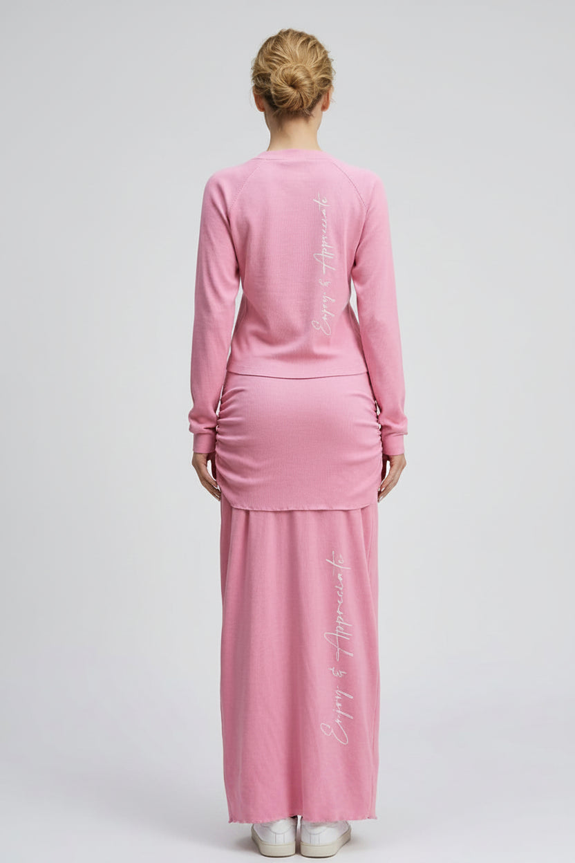 Woman wearing a pink long skirt and long sleeve top with text enjoy & apprecaite on a white background